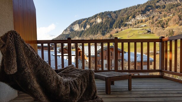 A wooden deck with a bench and a fur-covered item, overlooking a mountainous landscape with buildings. - '12 MN Megève Studio Comfort' with Mountain View (Flumet)