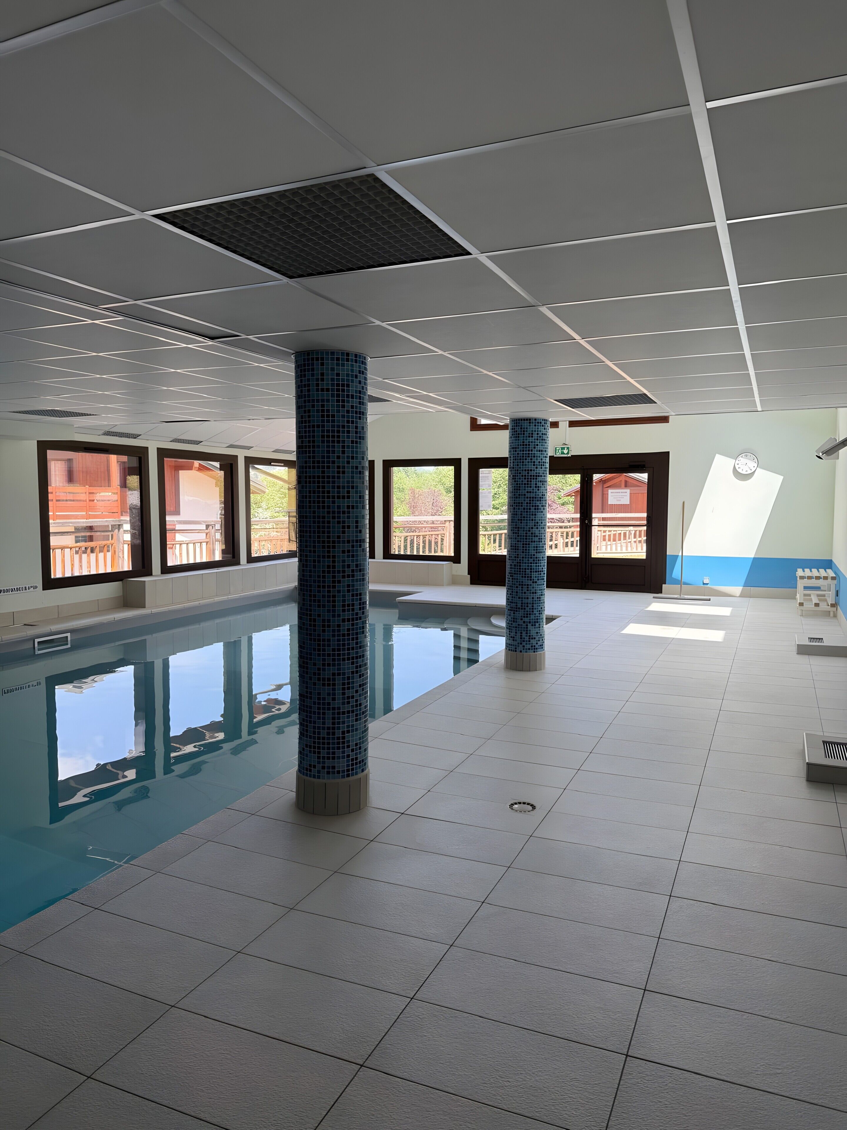 An indoor swimming pool with tiled floors and walls, large windows, and blue-tiled pillars.