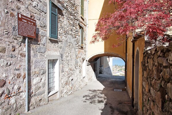 A narrow stone alleyway with a sign, a window with shutters, and a tree with red leaves.