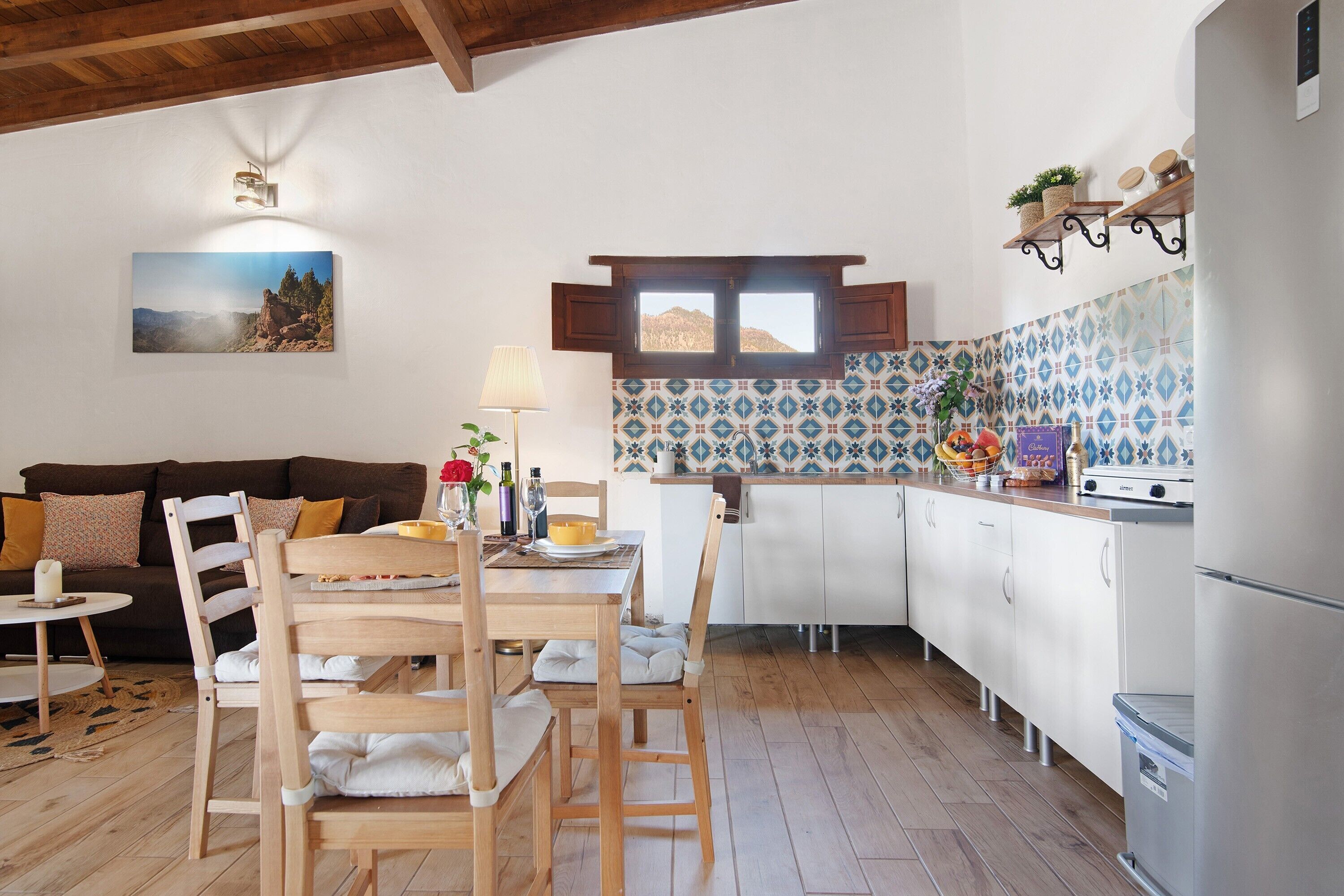 A modern kitchen with white cabinets, a wooden dining table, and a view of the mountains through a window.