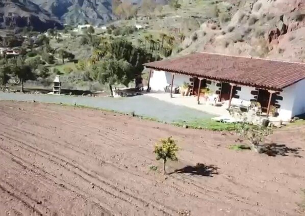 A rural house with a tiled roof, surrounded by a field and mountains.
