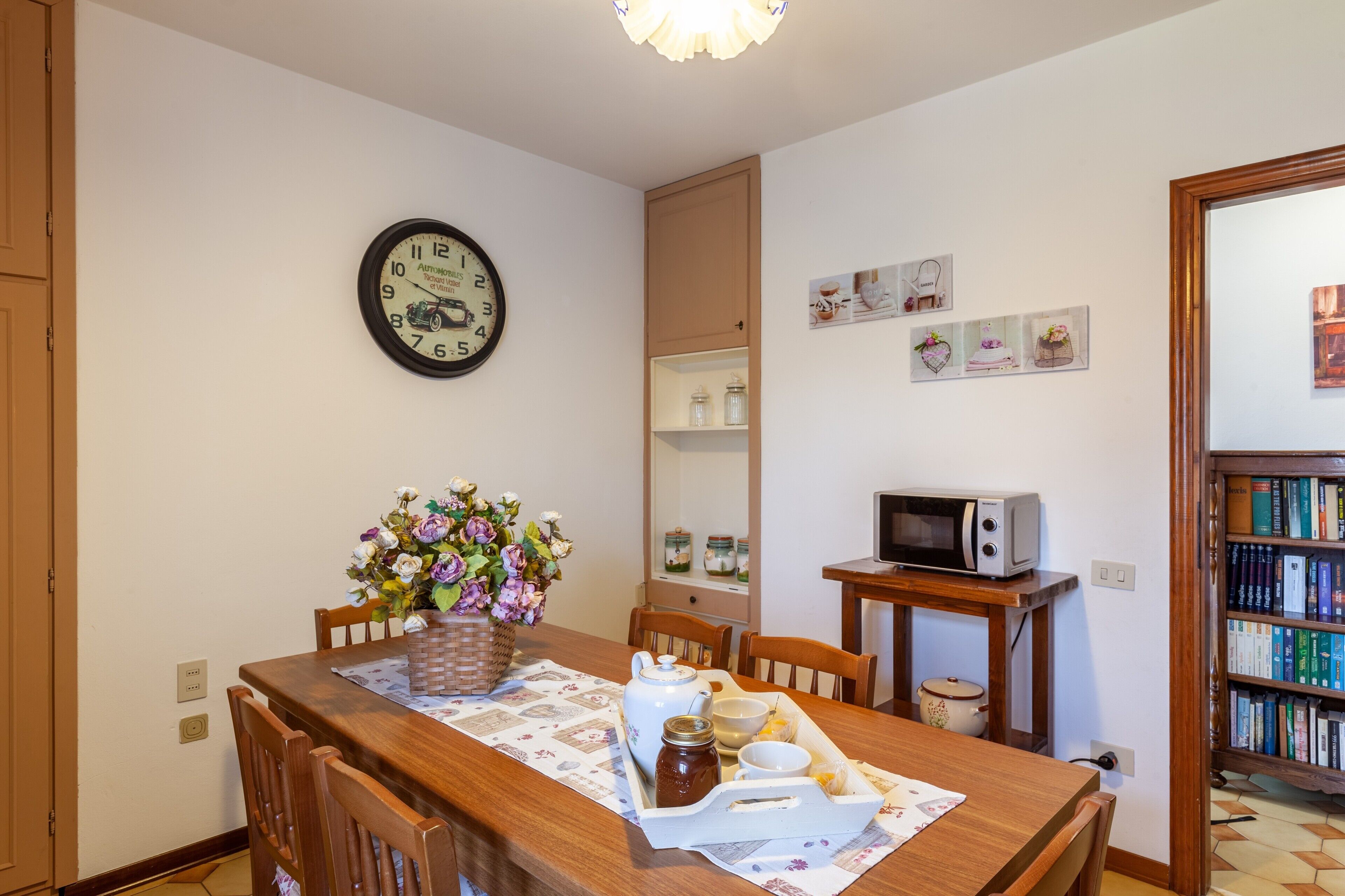 A dining area with a wooden table, chairs, a clock, a microwave, and a bookshelf.