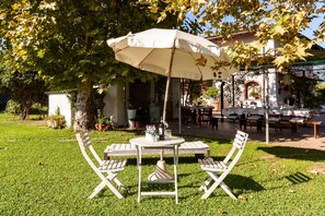Un espace extérieur aménagé avec une table et des chaises sous une marquise, entouré de verdure et bordé d’un bâtiment dont le toit est recouvert de tuiles.