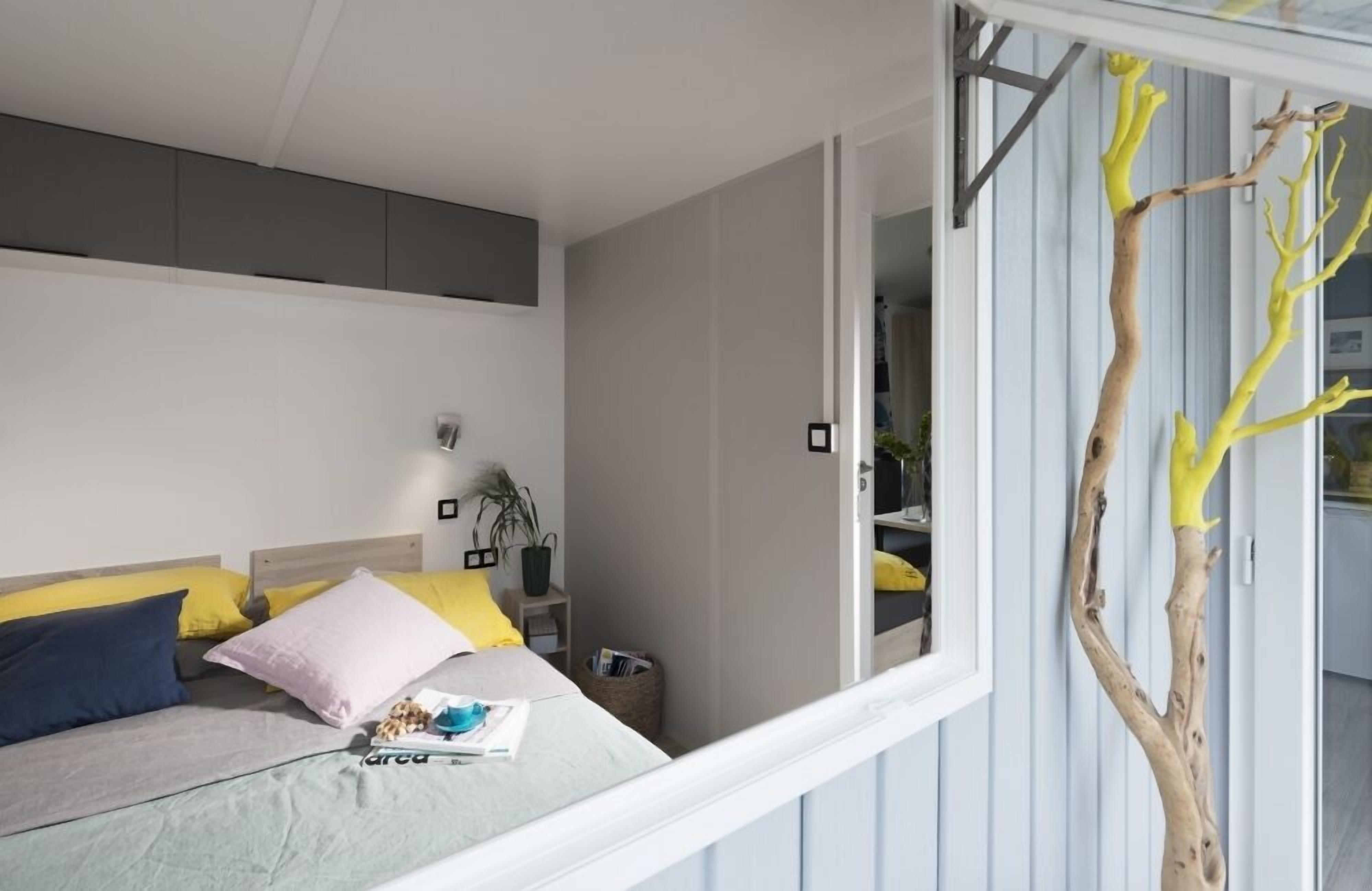 Un dormitorio con cama, almohadas, una mesita de noche, una lámpara y una ventana que da a un árbol.