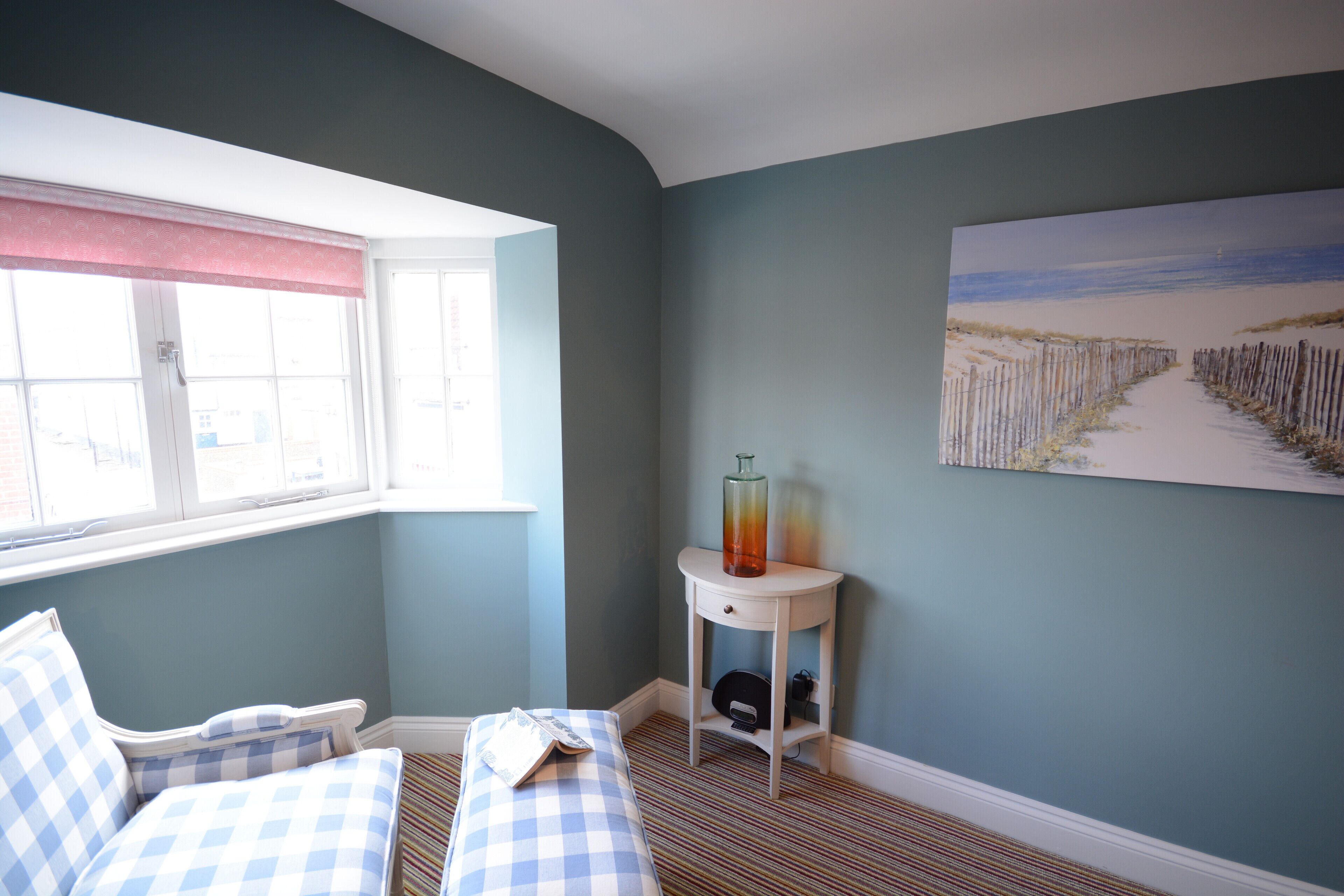 A bedroom with a checkered bed, a window with a view, a bedside table with a vase, and a painting on the wall.