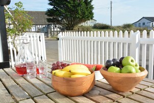 Una mesa de madera con cuencos de fruta y una jarra de bebida roja, frente a una valla de piquetes blancos con casas al fondo.