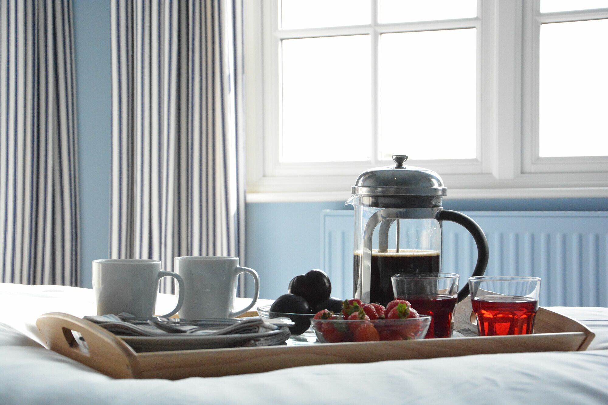 A breakfast tray with a French press, two cups, a glass, and a bowl of fruit on a bed.