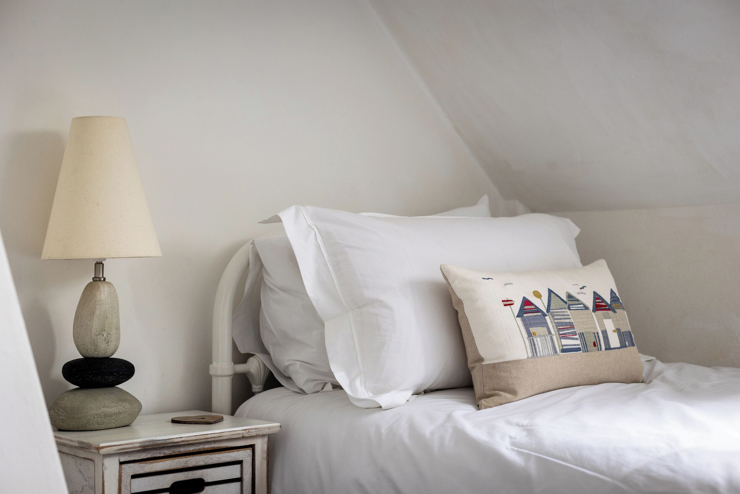 A neatly made bed with white linens and a decorative pillow, a bedside lamp, and a small stack of stones on a nightstand.