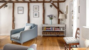 A cozy living room with exposed wooden beams, a blue armchair, a wooden bench, a white console table with books, and a vase with flowers. - 7 Church Street, Southwold (Reydon)