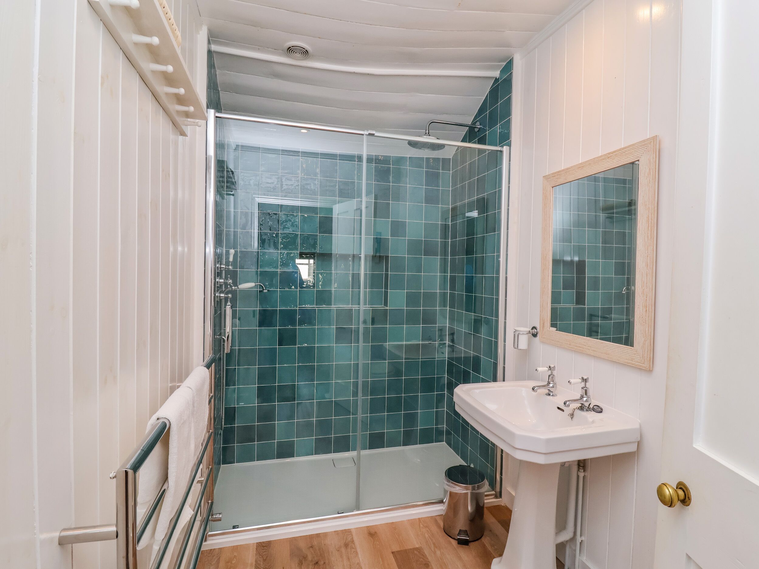 A bathroom with a glass shower enclosure, a white sink with a mirror, and a wooden floor.