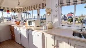 A kitchen with white cabinets, a microwave, a kettle, and a window with a view of houses and trees. - Valetta, Thorpeness (Leiston)