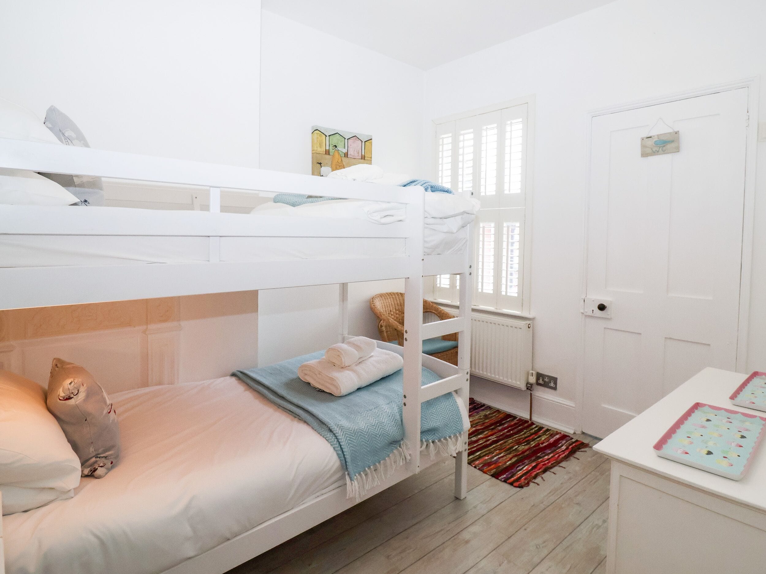 A bunk bed room with a white door, a small table with a colorful rug, and a window with blinds.