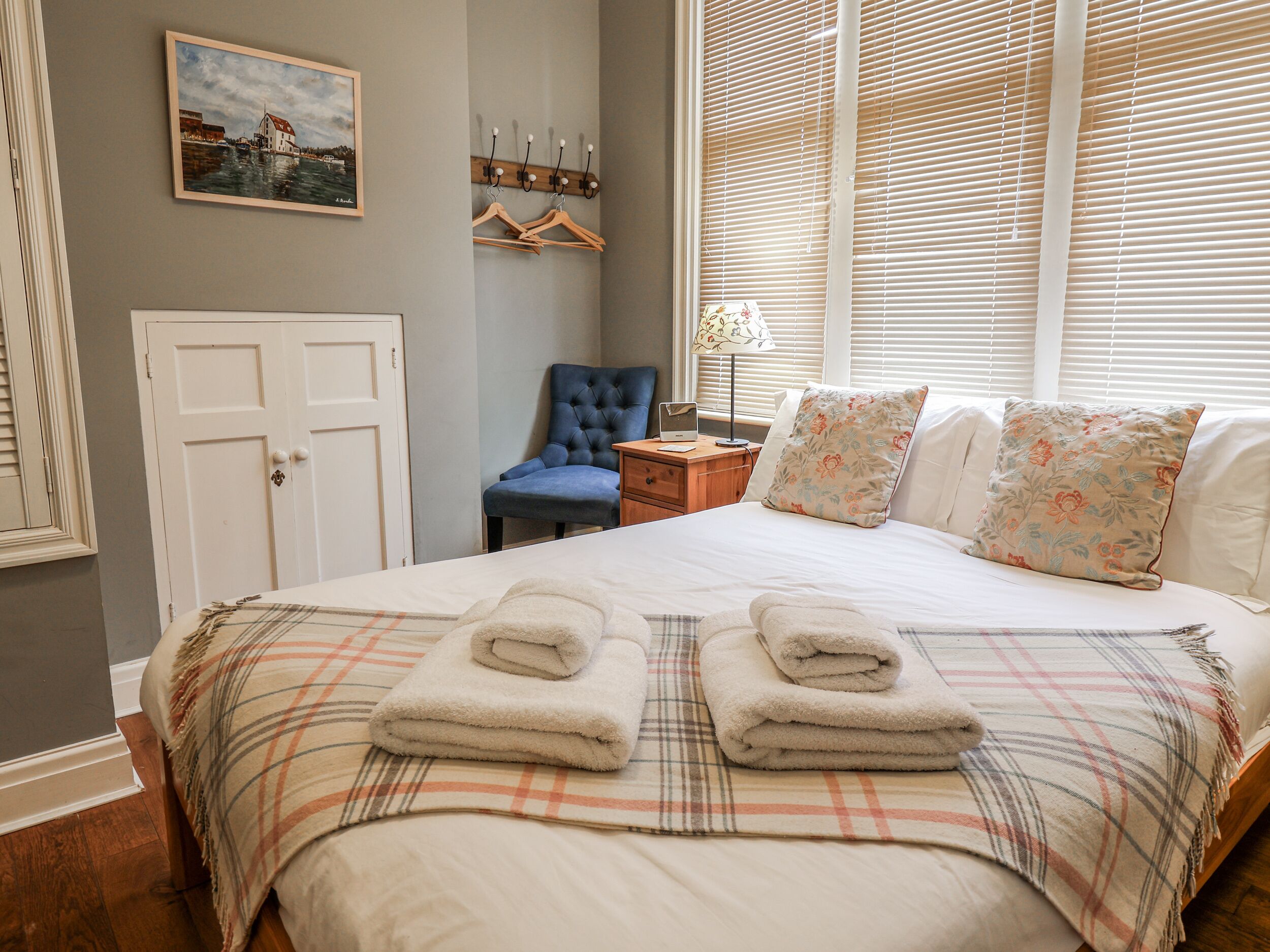 Un lit bien fait, recouvert d’une couverture à carreaux, avec deux serviettes pliées, un fauteuil bleu, une table de chevet en bois avec une lampe, un porte-manteau avec des cintres et un tableau encadré accroché au mur.