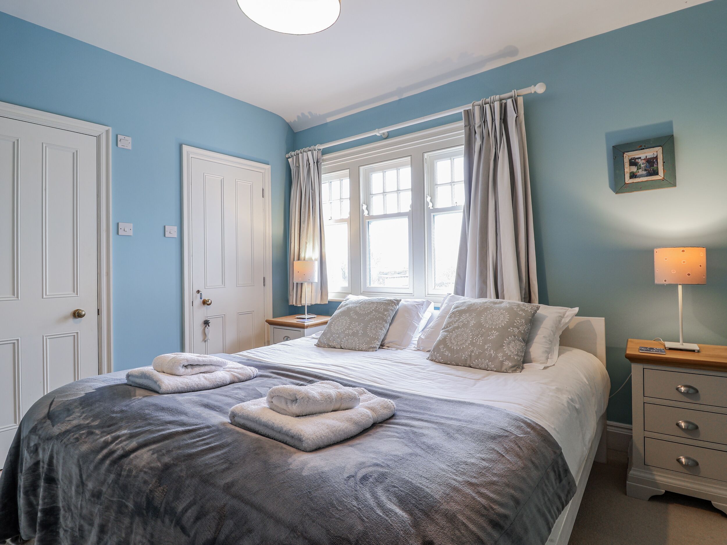 A bedroom with a bed, two bedside tables, a nightstand, a window with curtains, and a wall-mounted picture.