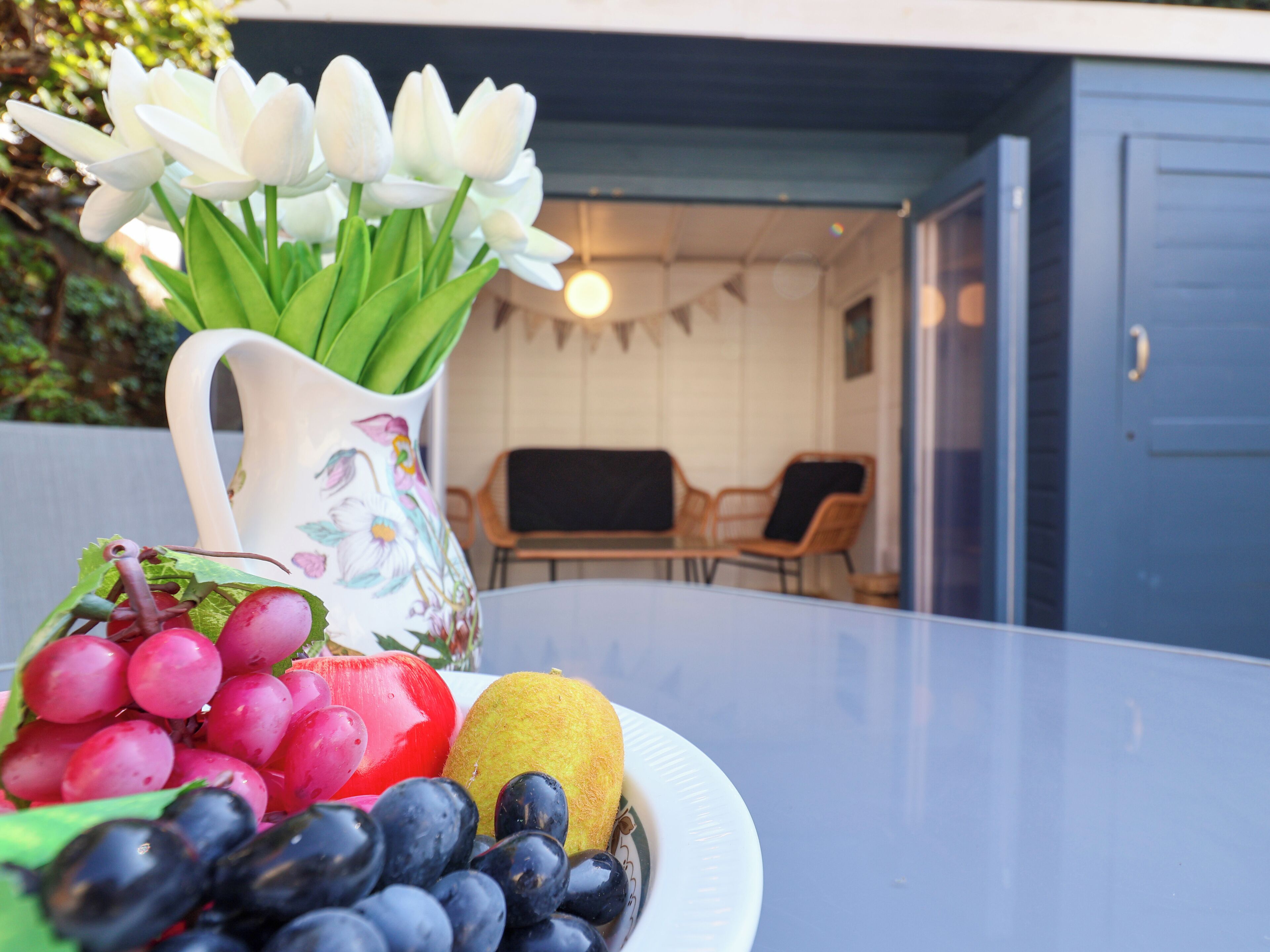 A table with a fruit and flower arrangement, a pitcher, and a view of an outdoor seating area.