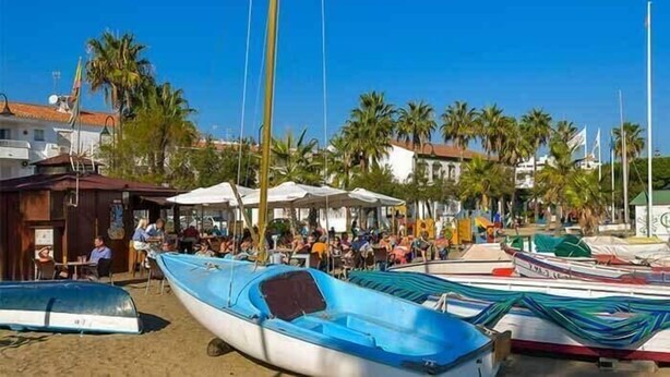 A beachside area with boats, palm trees, and a building with a red roof.