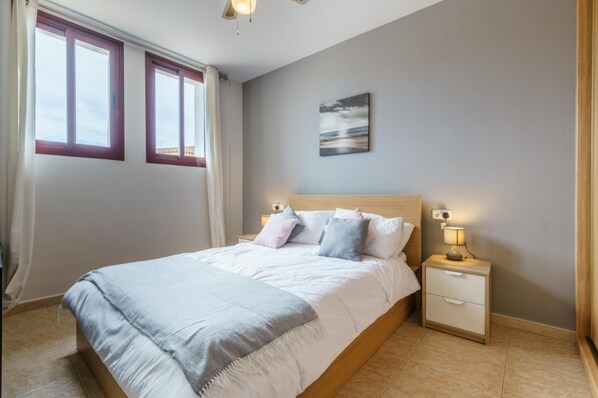 Un dormitorio con cama, mesitas de noche, un cuadro y una ventana con cortinas.