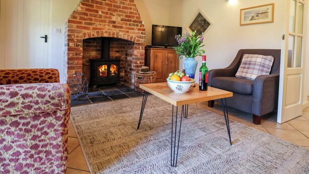 A cozy living room with a brick fireplace, a wooden table with a bowl of fruit and a bottle, a grey sofa, and a patterned armchair.