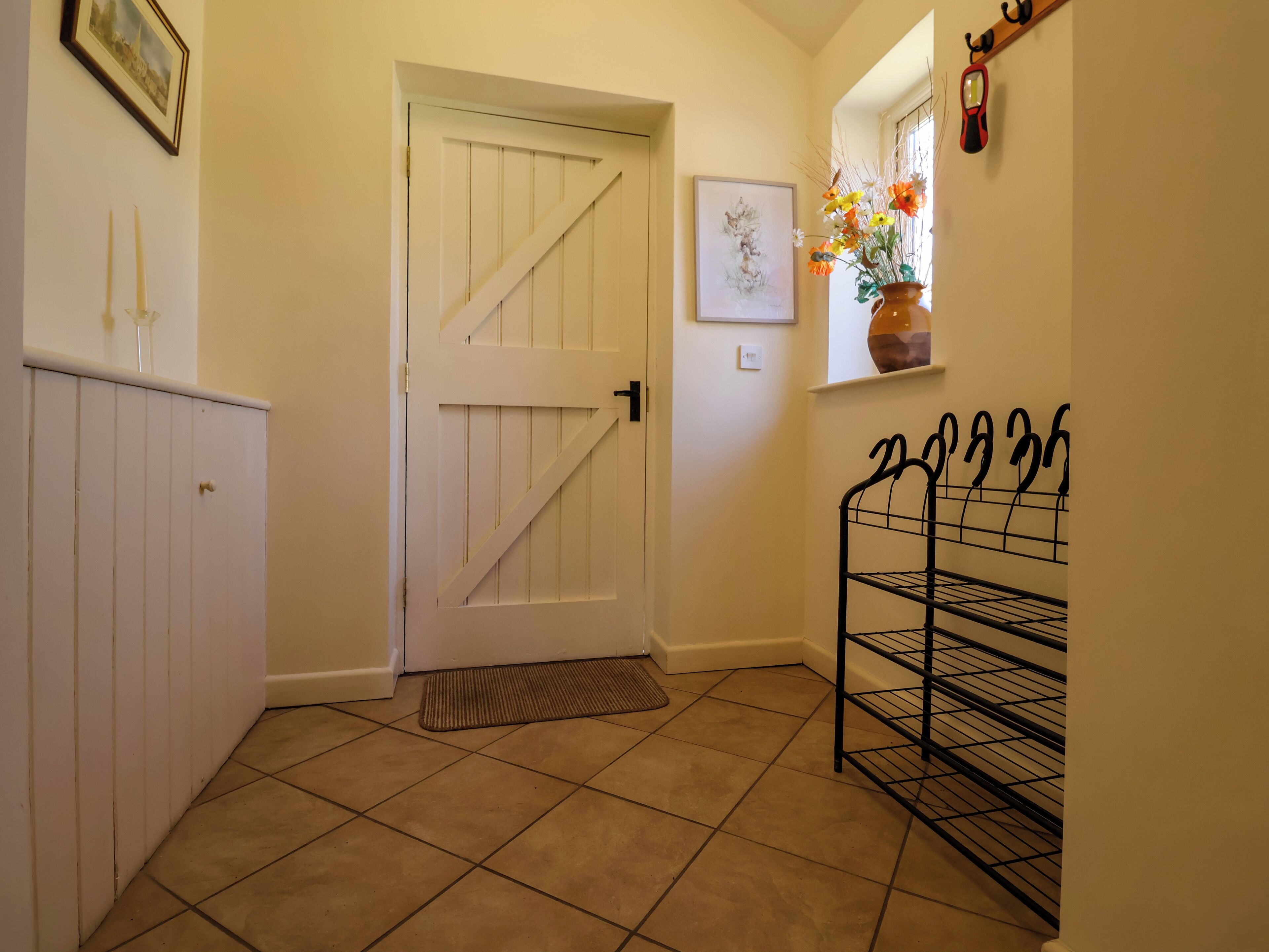 A hallway with a white door, a black metal rack, a tiled floor, and a small window with a vase of flowers.