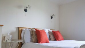Una cama con ropa blanca y varias almohadas, una mesita de noche con lámpara, y un estante blanco pequeño con una caja de pañuelos.