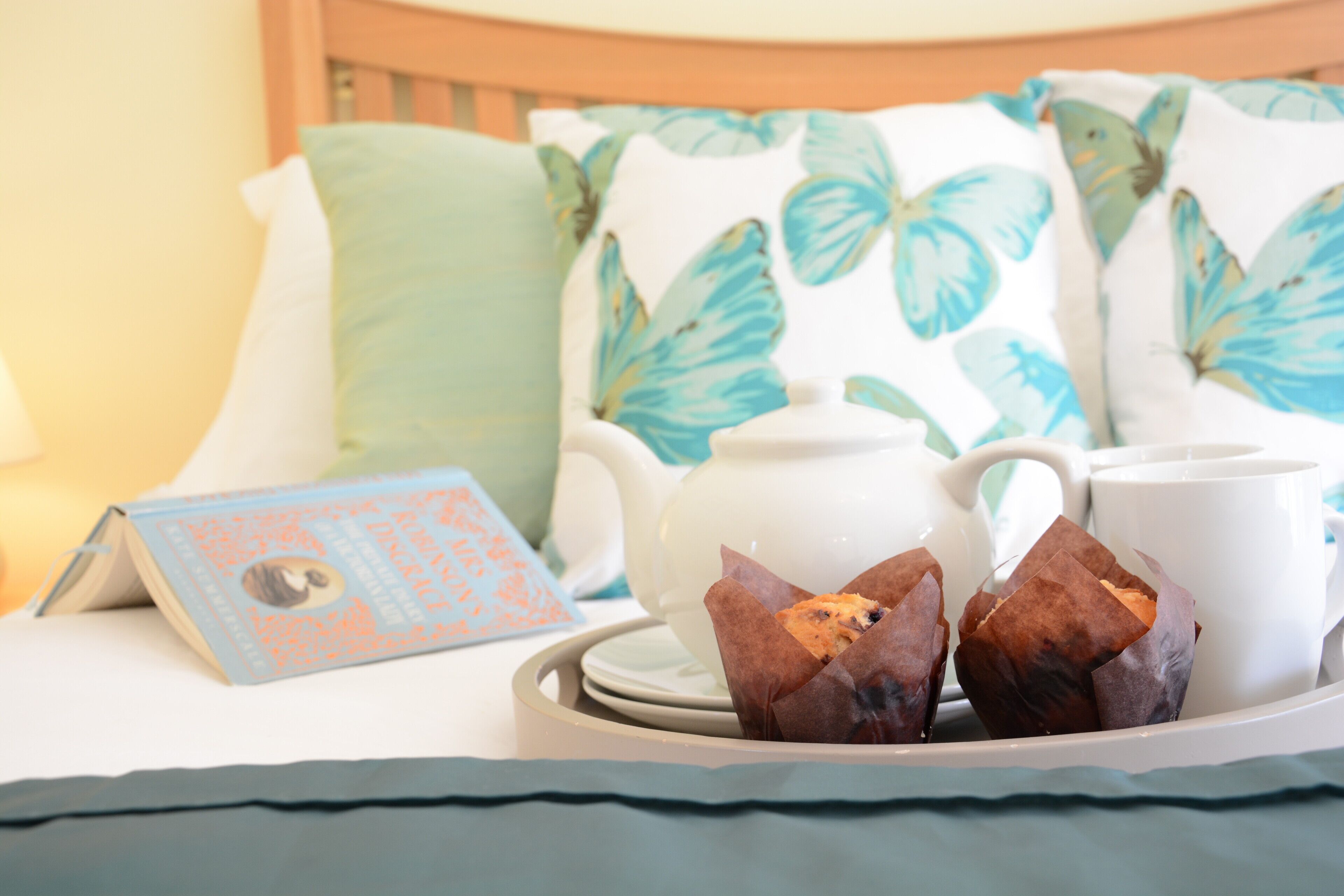 Una bandeja con dos muffins sobre una cama, junto con un libro y una tetera.