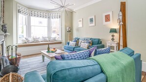 A living room with a blue sofa, a wooden coffee table, and a window seat with patterned cushions. - Heron House, Southwold (Reydon)