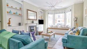 A living room with teal sofas, a fireplace, a large mirror, and a view of the street through large windows. - Heron House, Southwold (Reydon)