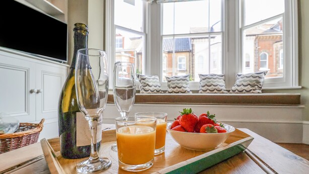 A breakfast tray with a bottle, glasses, and a bowl of strawberries on a wooden table by a window with a view of buildings. - Heron House, Southwold (Reydon)