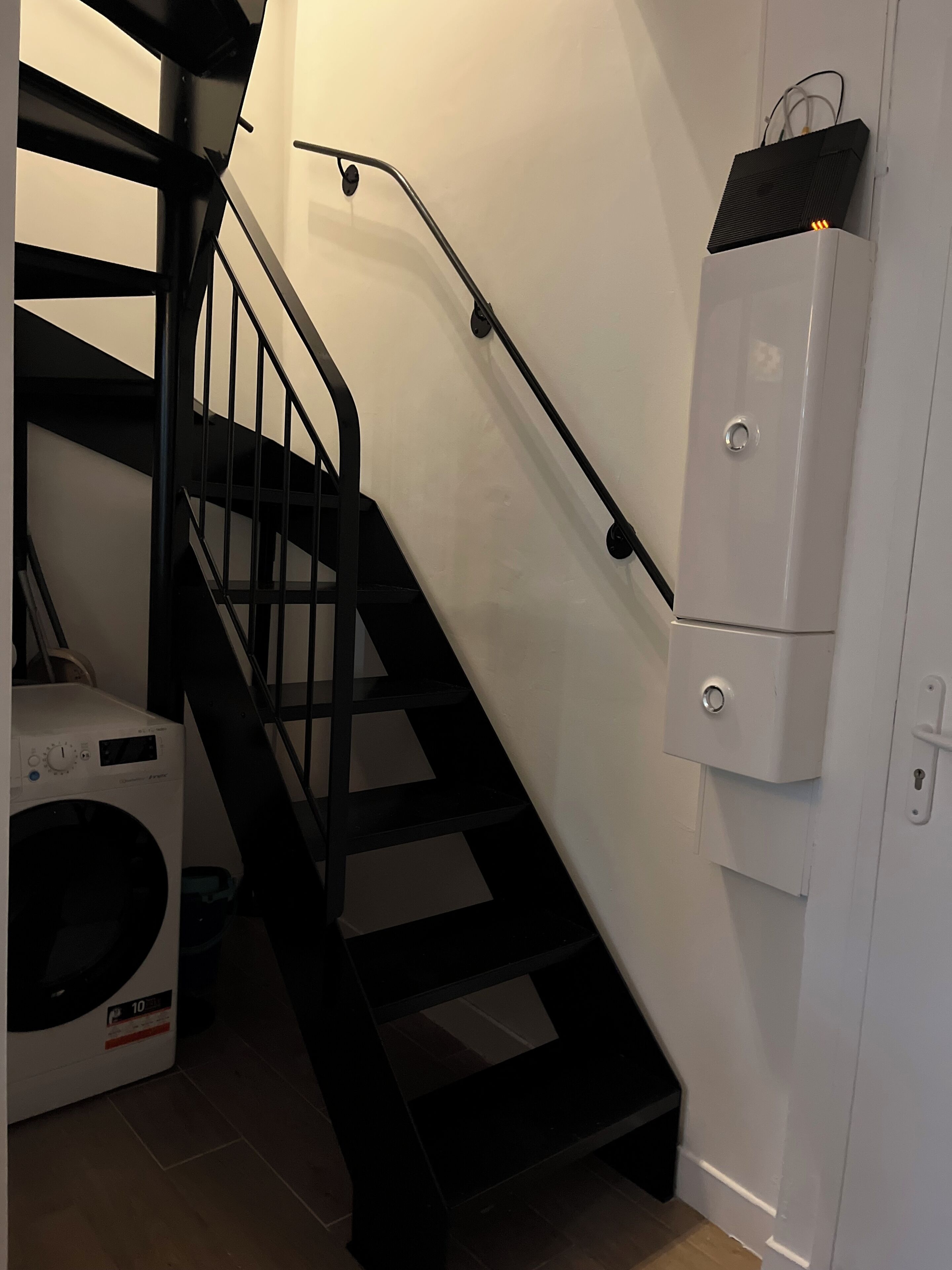 Een trap met een zwarte leuning, een witte wasmachine en een witte kast met een zwarte speaker erop.