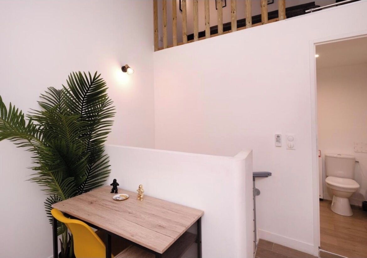 Une petite pièce avec une table en bois, une chaise jaune, une plante en pot et un mur blanc.