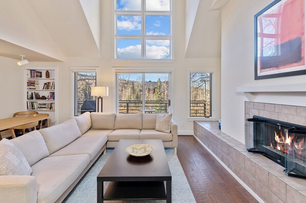 A spacious living room with a high ceiling, a fireplace, a sofa, a coffee table, and a bookshelf.
