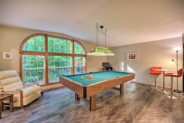 Une table de billard dans une pièce dotée d’une grande fenêtre, d’une chaise de billard et de deux tabourets de bar.