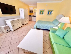 Ein Zimmer mit einem Bett, einem grünen Sofa, einem weißen Tisch, einem an der Wand befestigten Fernseher und einer Küchenzeile im Hintergrund.