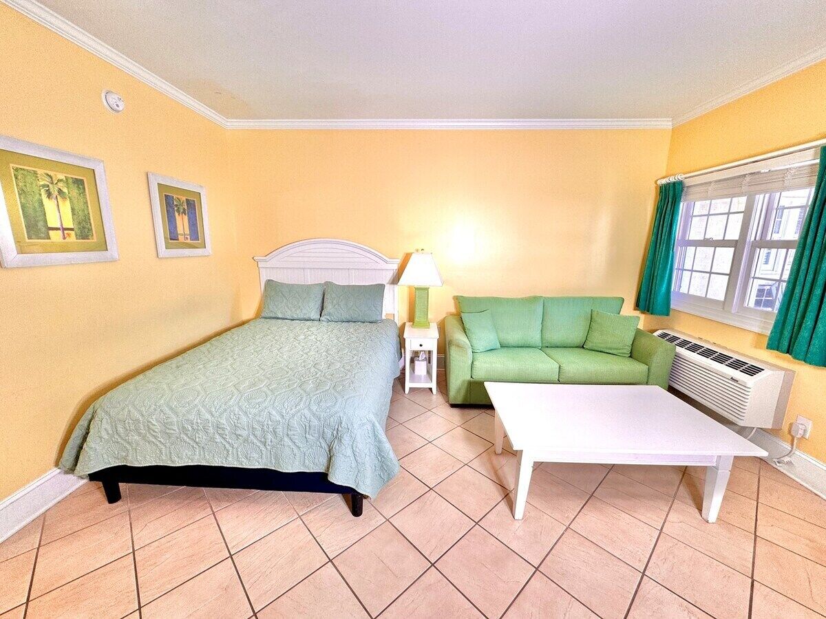A room with a bed, a green sofa, a white table, a lamp, and a window with curtains.