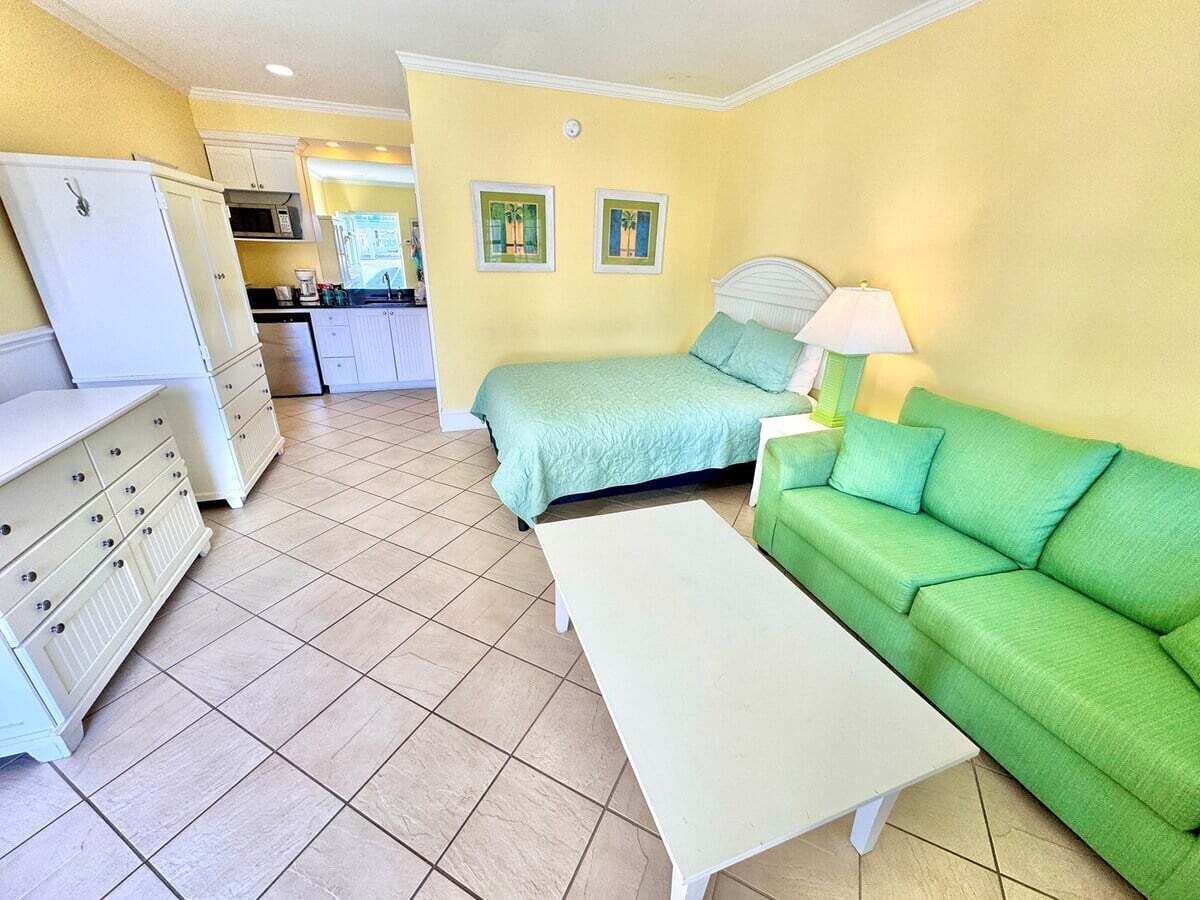 Una habitación con una cama, un sofá verde, una mesa blanca, una zona de cocina y un refrigerador blanco.