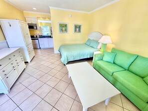 Ein Zimmer mit einem Bett, einem grünen Sofa, einem weißen Tisch, einer Küchenzeile und einem weißen Kühlschrank.