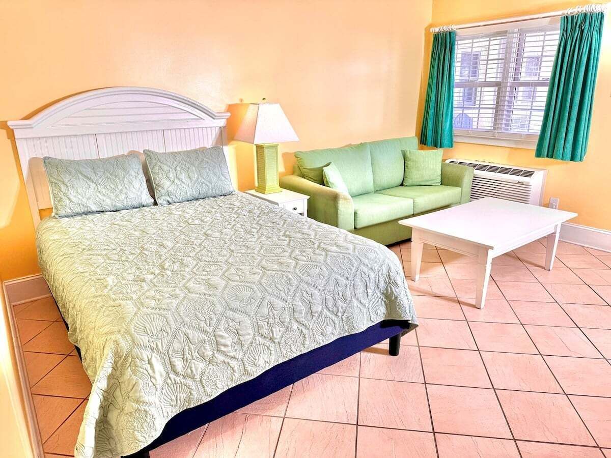Une pièce avec un lit, un canapé vert, une table blanche et une fenêtre avec des rideaux.