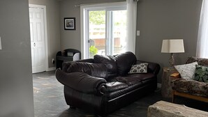 A living room with a black leather sofa, a wooden coffee table, and a patterned armchair. There is a door leading to another room and a window with curtains.