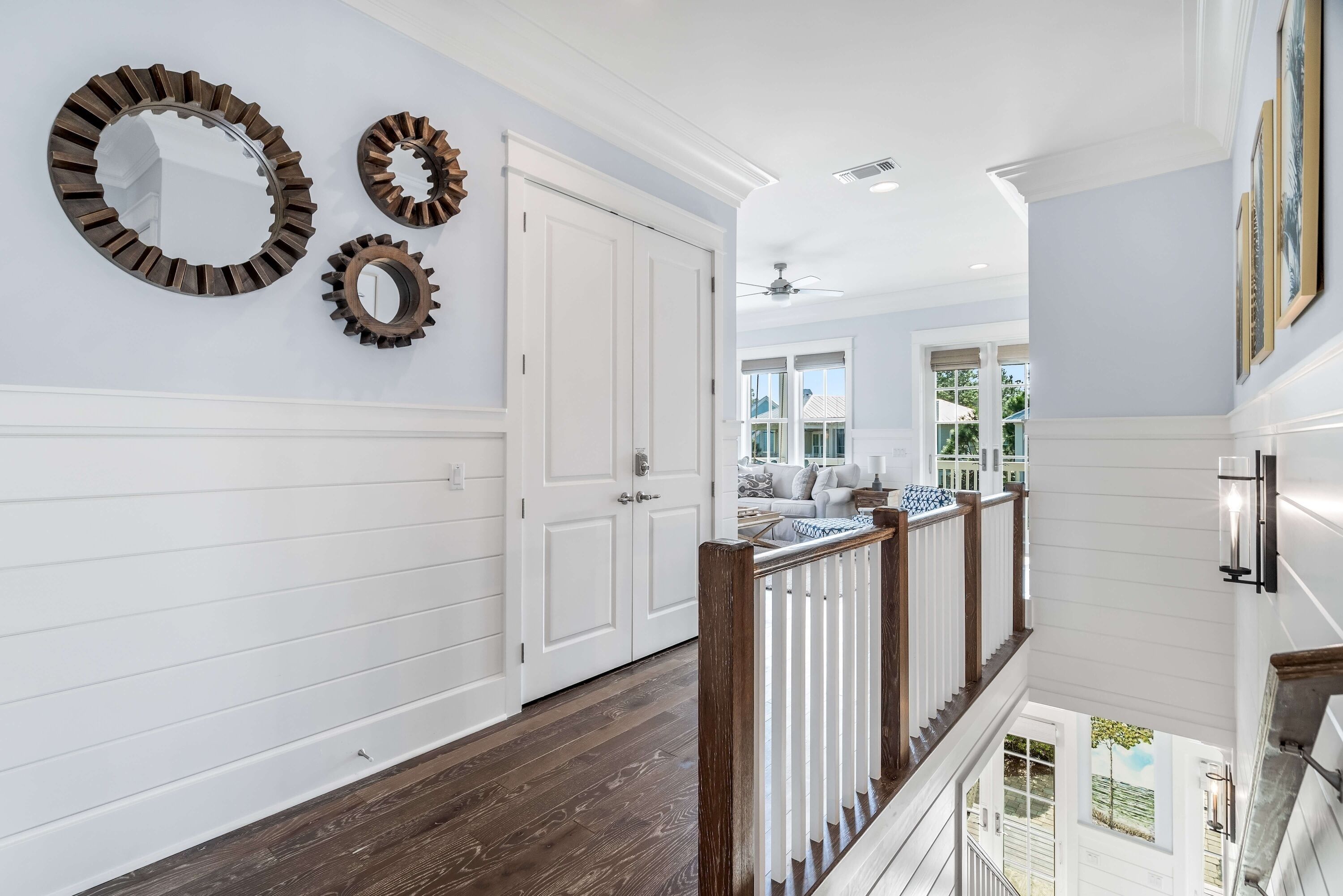 A hallway with a wooden floor, white paneling, and a white door. There are three wall-mounted mirrors and a ceiling fan.