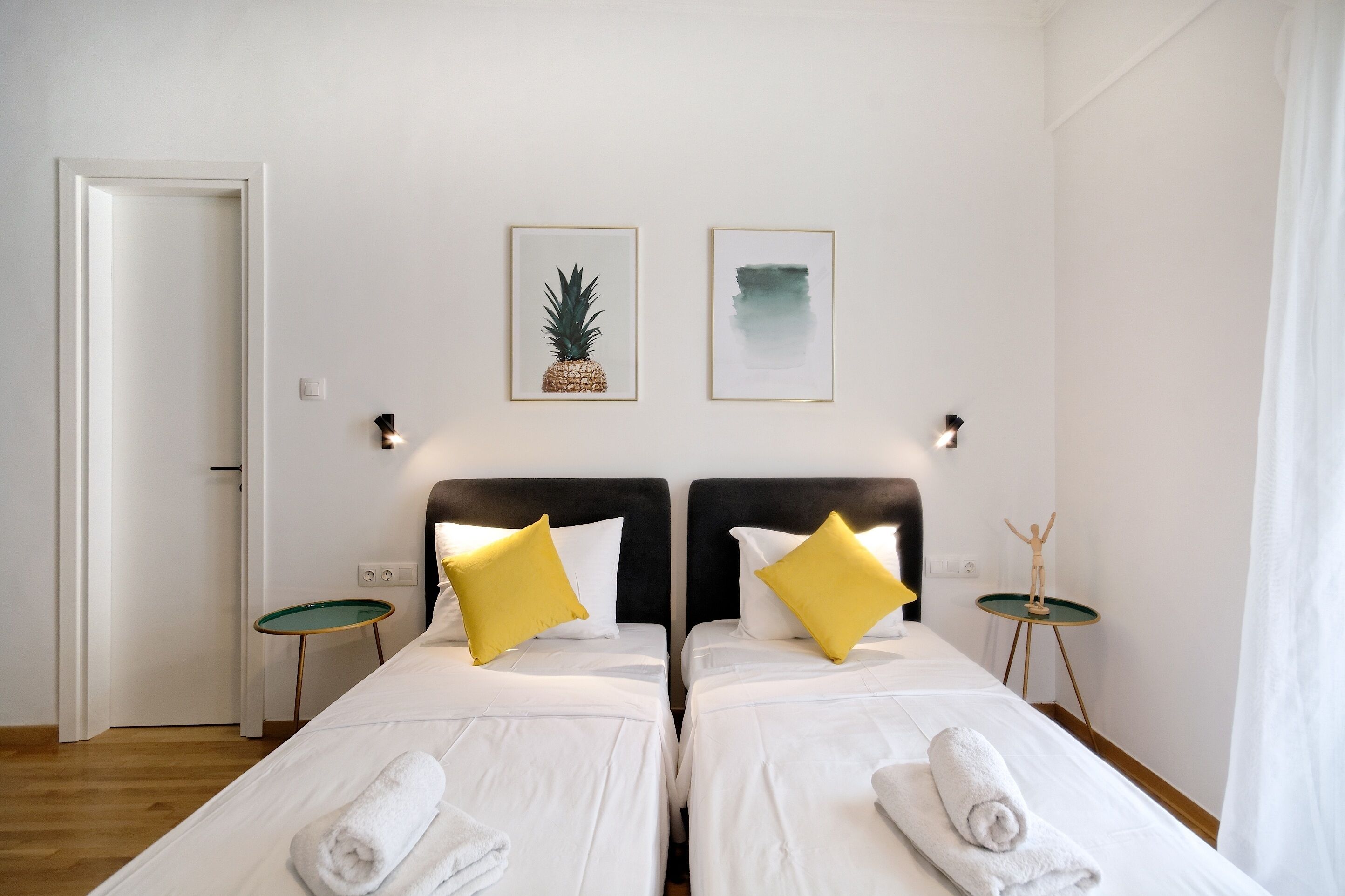 Habitación de hotel con dos camas, ropa de cama blanca, almohadas amarillas, una mesita con una estatua y dos cuadros en la pared.