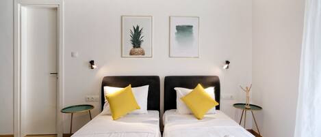 Habitación de hotel con dos camas, ropa de cama blanca, almohadas amarillas, una mesita con una estatua y dos cuadros en la pared.
