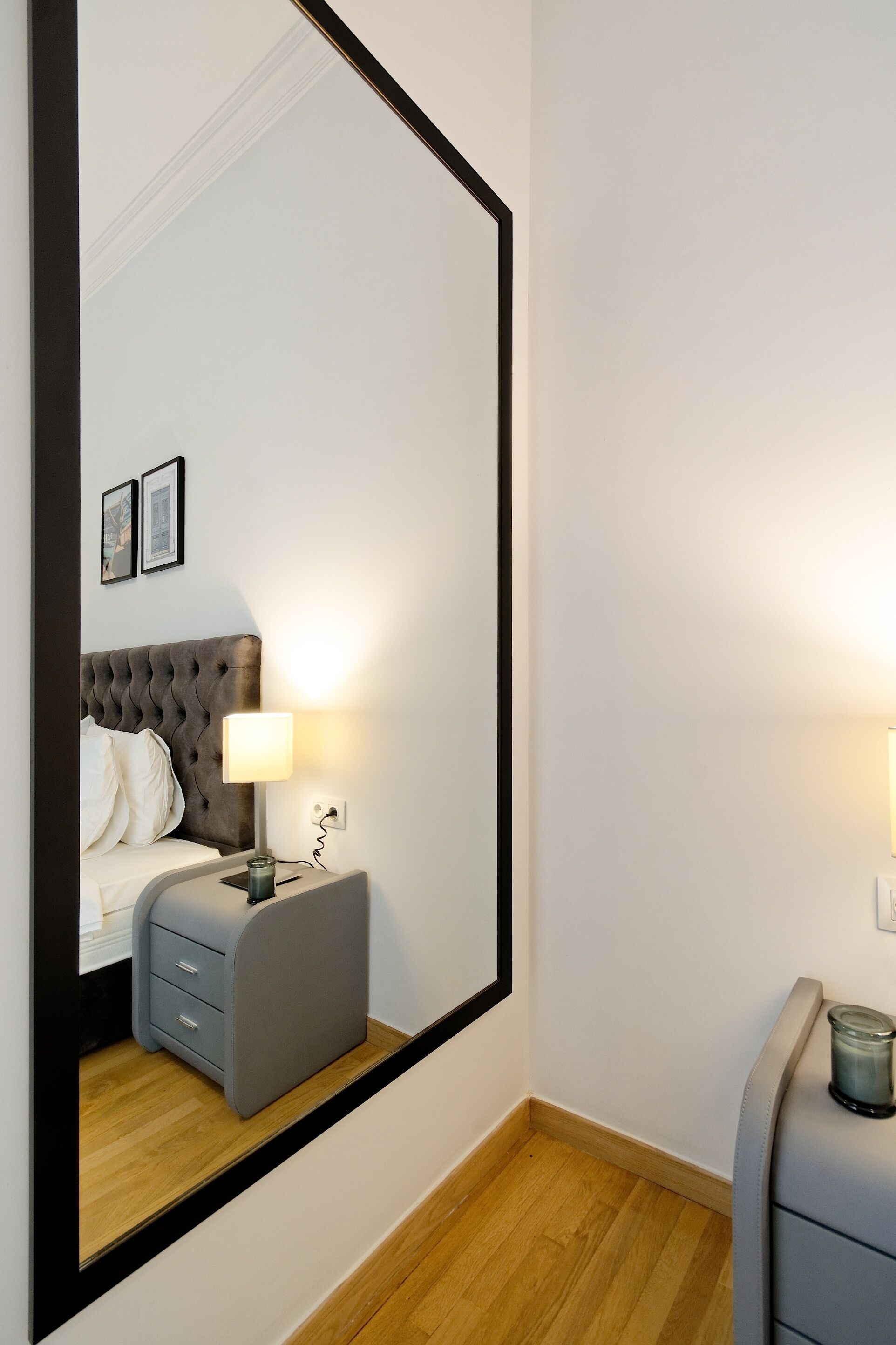 A bedroom with a large mirror, a bed with a tufted headboard, a bedside table with a lamp, and a small drawer unit.