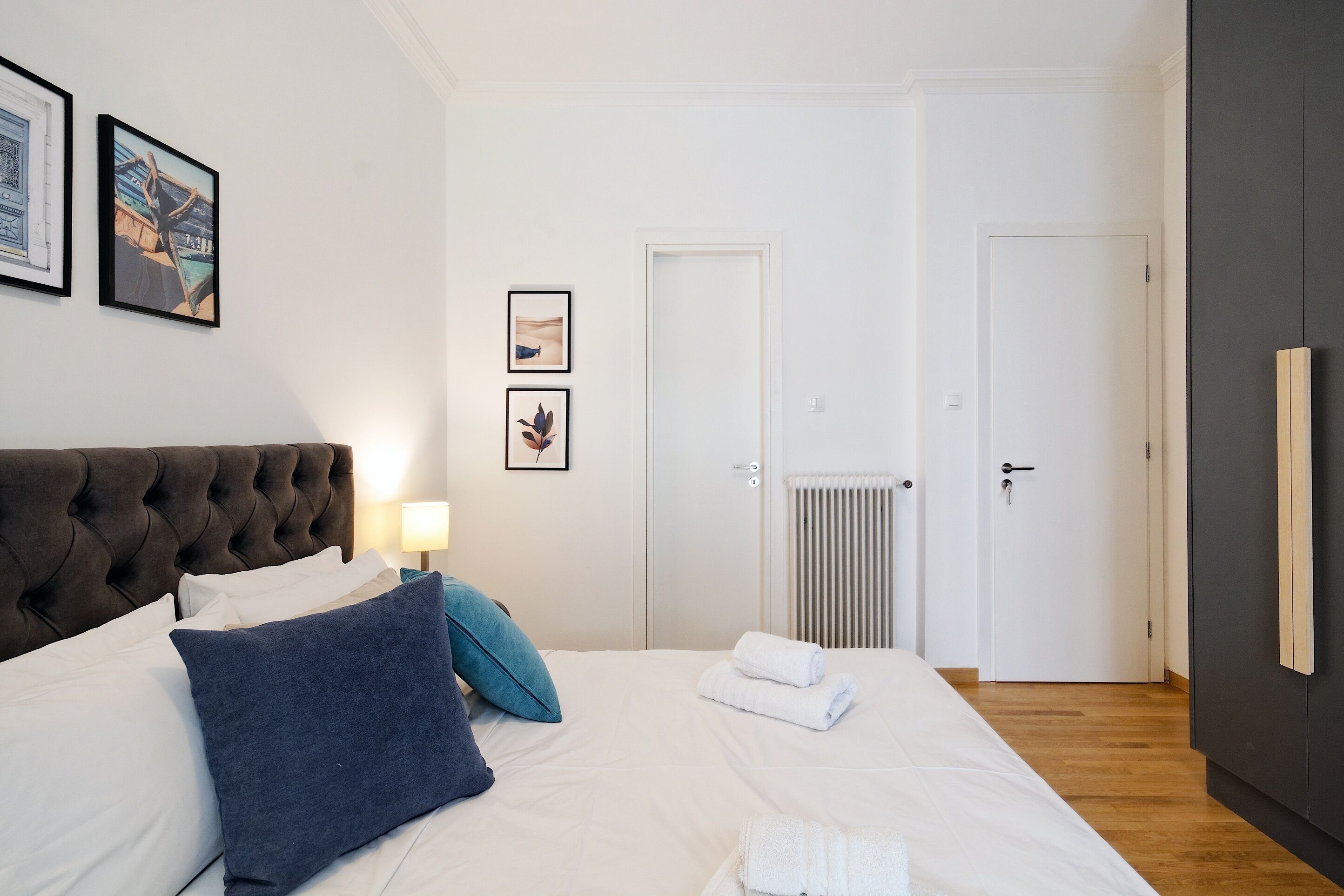 A neatly made bed with white linens and blue pillows, a wall with framed artwork, and a door leading to another room.