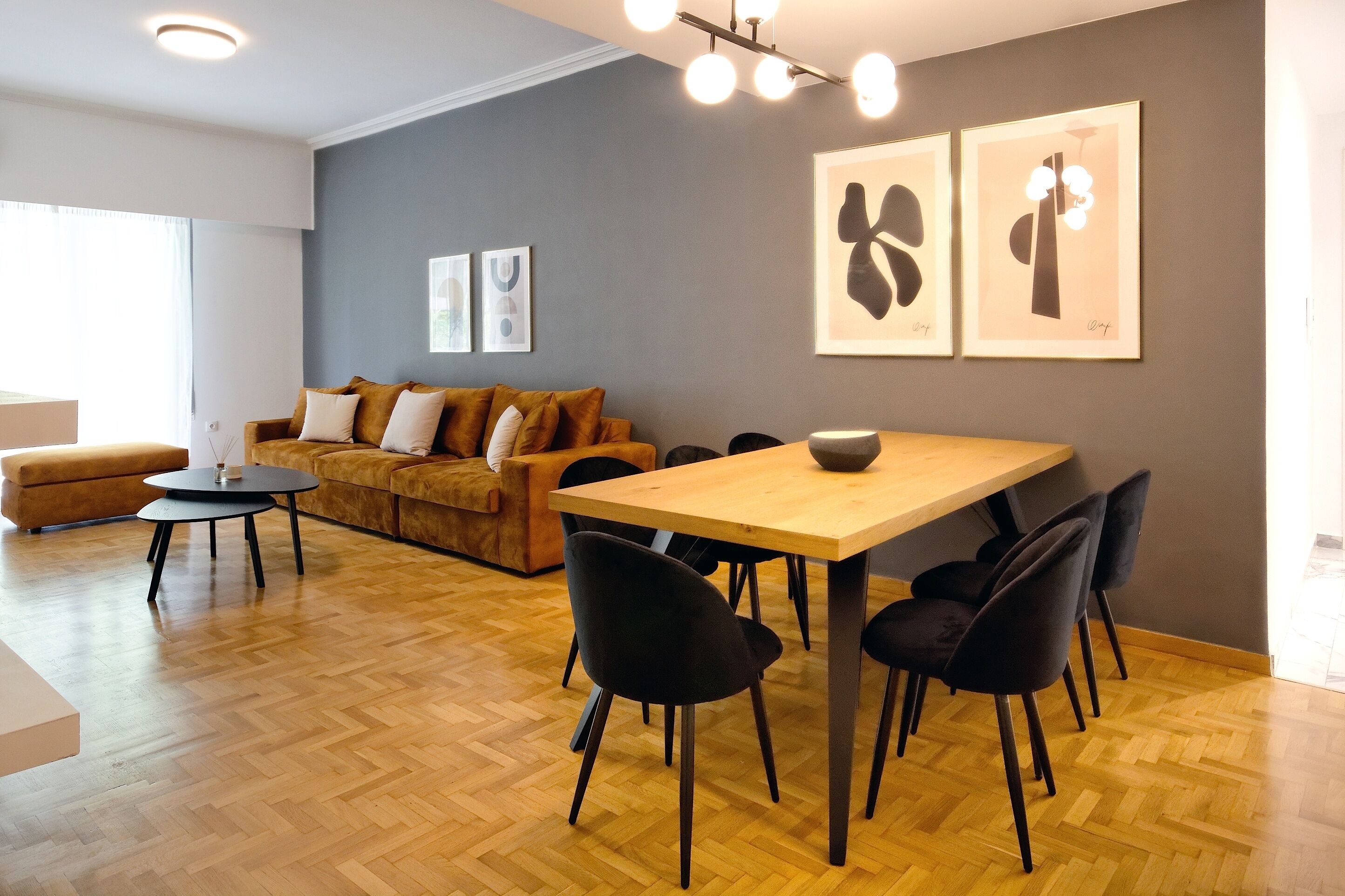 A modern living room with a wooden dining table, black chairs, a sofa, and a coffee table. There are framed artworks on the wall and a window providing natural light.