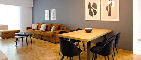 A modern living room with a wooden dining table, black chairs, a sofa, and a coffee table. There are framed artworks on the wall and a window providing natural light.