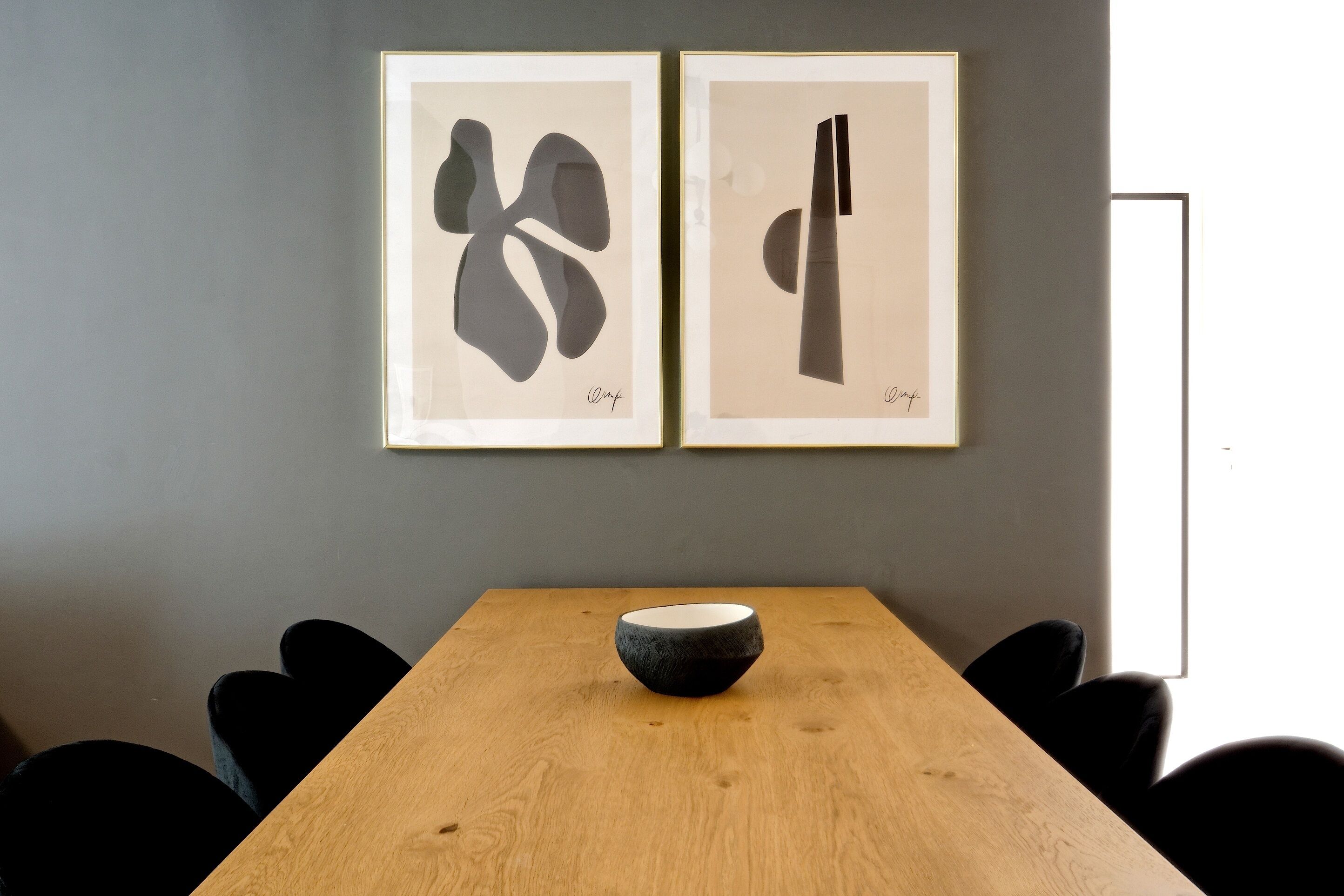 A wooden table with a black bowl in the center, flanked by black chairs, against a grey wall with two framed abstract artworks.