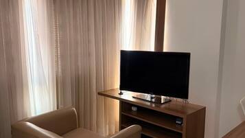 A hotel room with a flat-screen TV on a wooden stand, a leather armchair, a bed with a striped bedspread, and a window with sheer curtains.