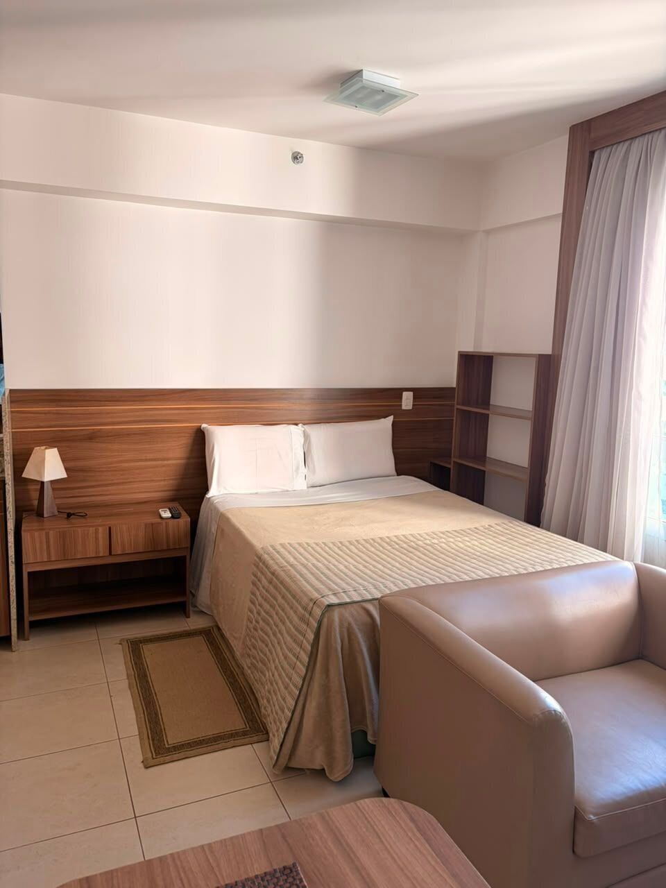 Una habitación de hotel con cama, mesita de noche, lámpara, silla y ventana con cortinas.