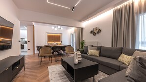 A modern living room with a sofa, coffee table, and TV. There is a dining area with chairs and a table in the background. - Phaedrus Living: Luxury Penthouse Ploutarchou (Athens)
