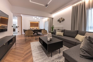 A modern living room with a sofa, coffee table, and TV. There is a dining area with chairs and a table in the background.